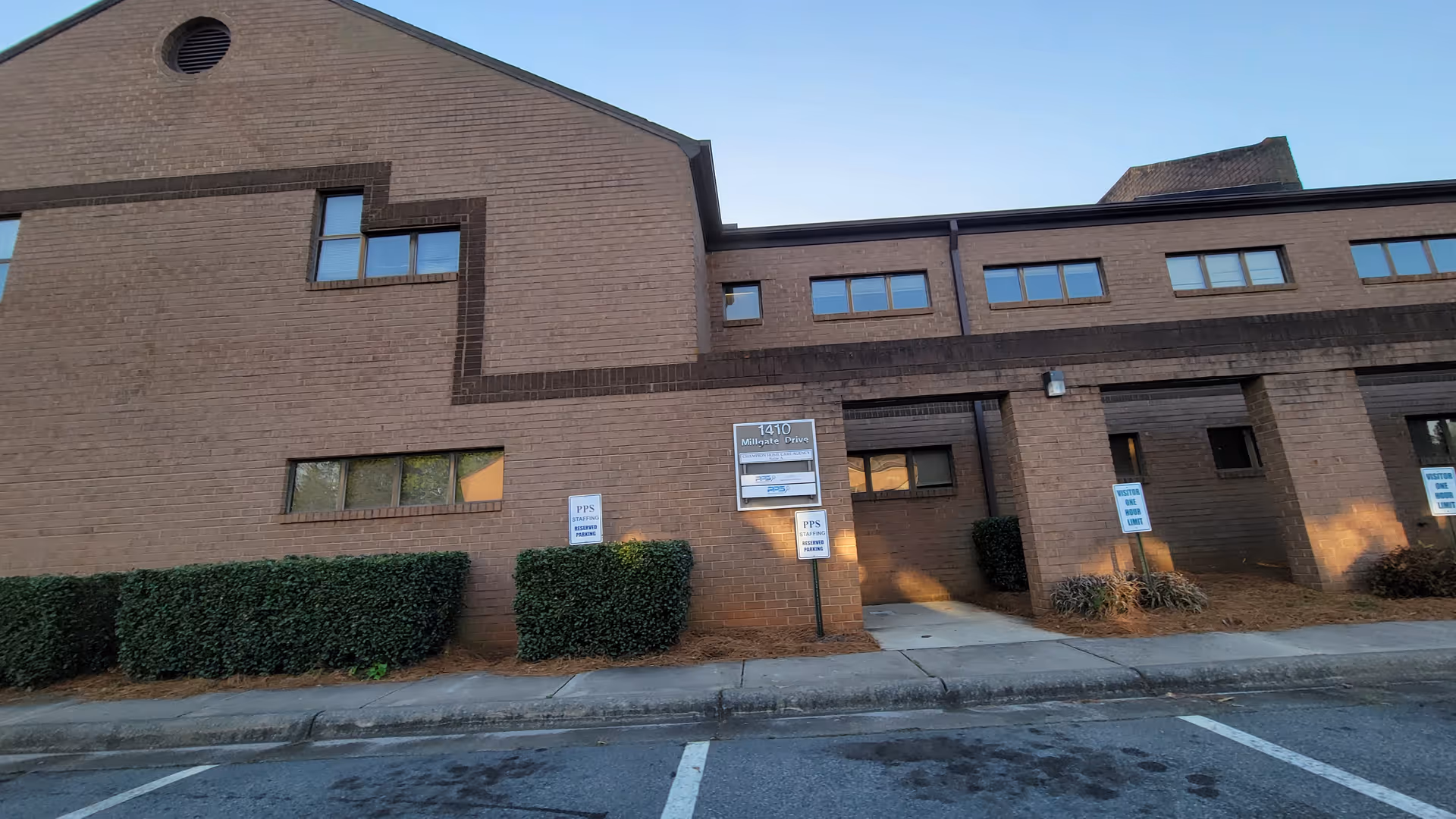 Brick senior living facility exterior with entryway, signage, parking, and trimmed hedges.
