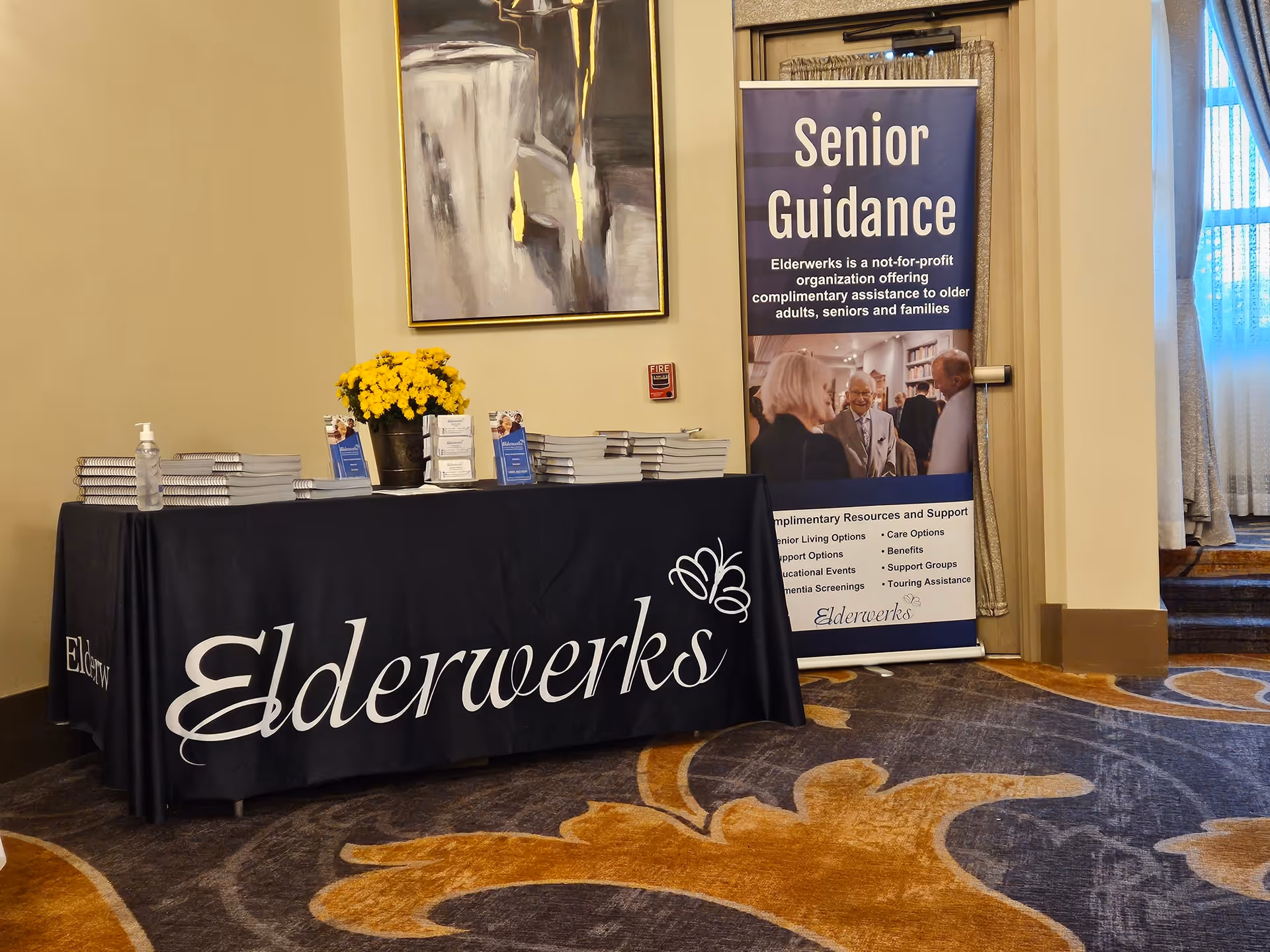 Indoor reception table with Elderwerks banner, brochure stacks, and yellow flowers.