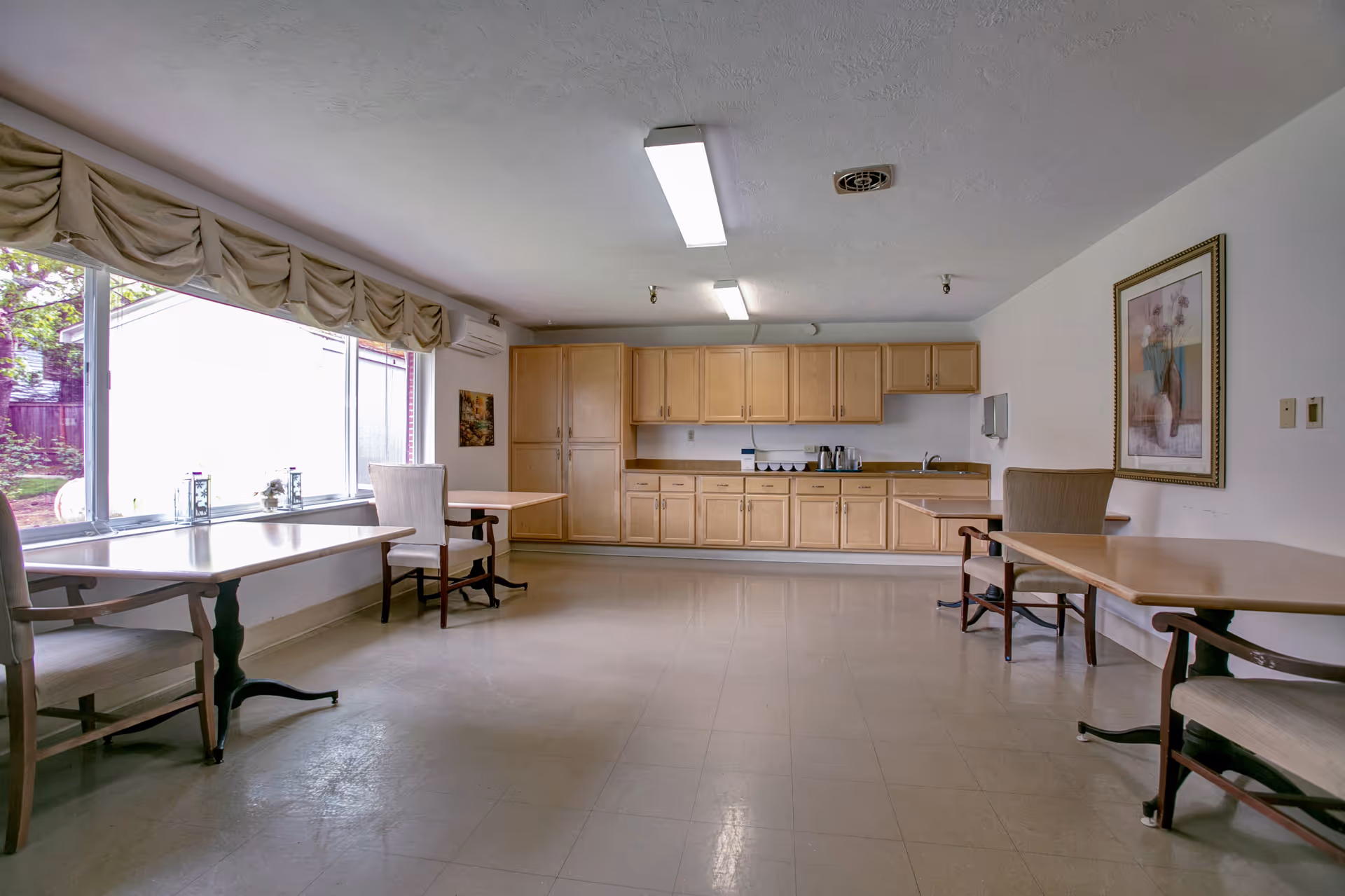 A spacious senior living dining area with a long kitchenette along the back wall and tables.