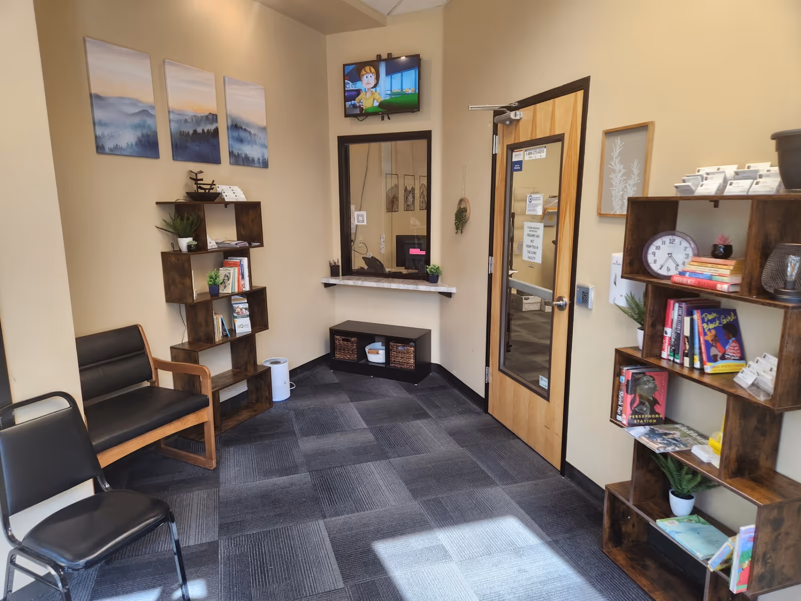 Beige-walled waiting area with black chairs, wooden shelves, TV, and doorway.