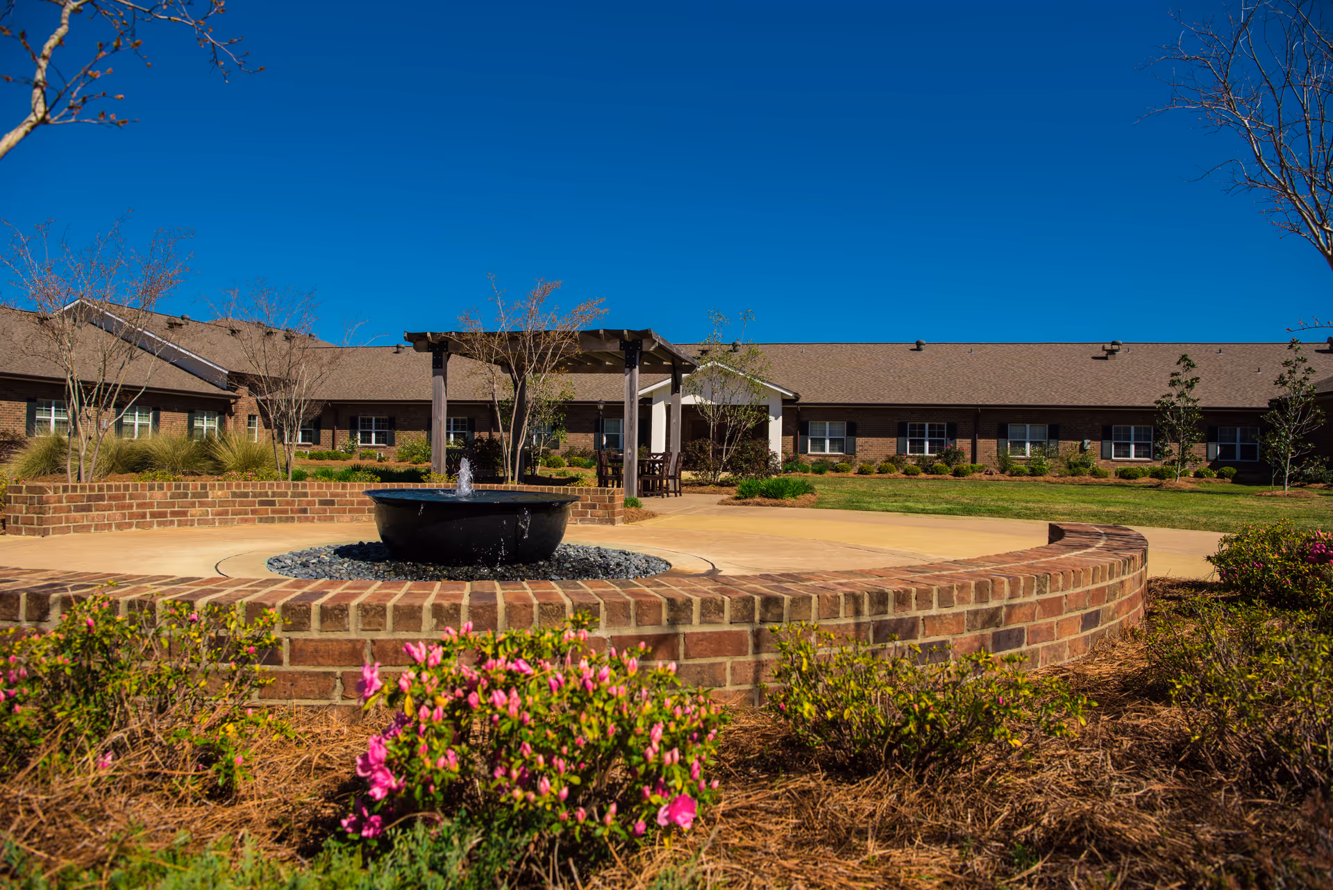 Brick courtyard with a central fountain and flowerbeds in front of a single-story rehabilitation center building under a clear blue sky.