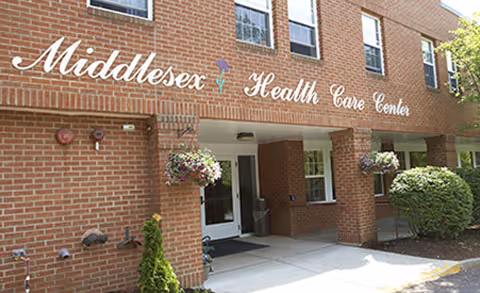Exterior view of the Middlesex Health Care Center building entrance with brick walls, windows, hanging flower baskets, and a small shrub near the doorway.