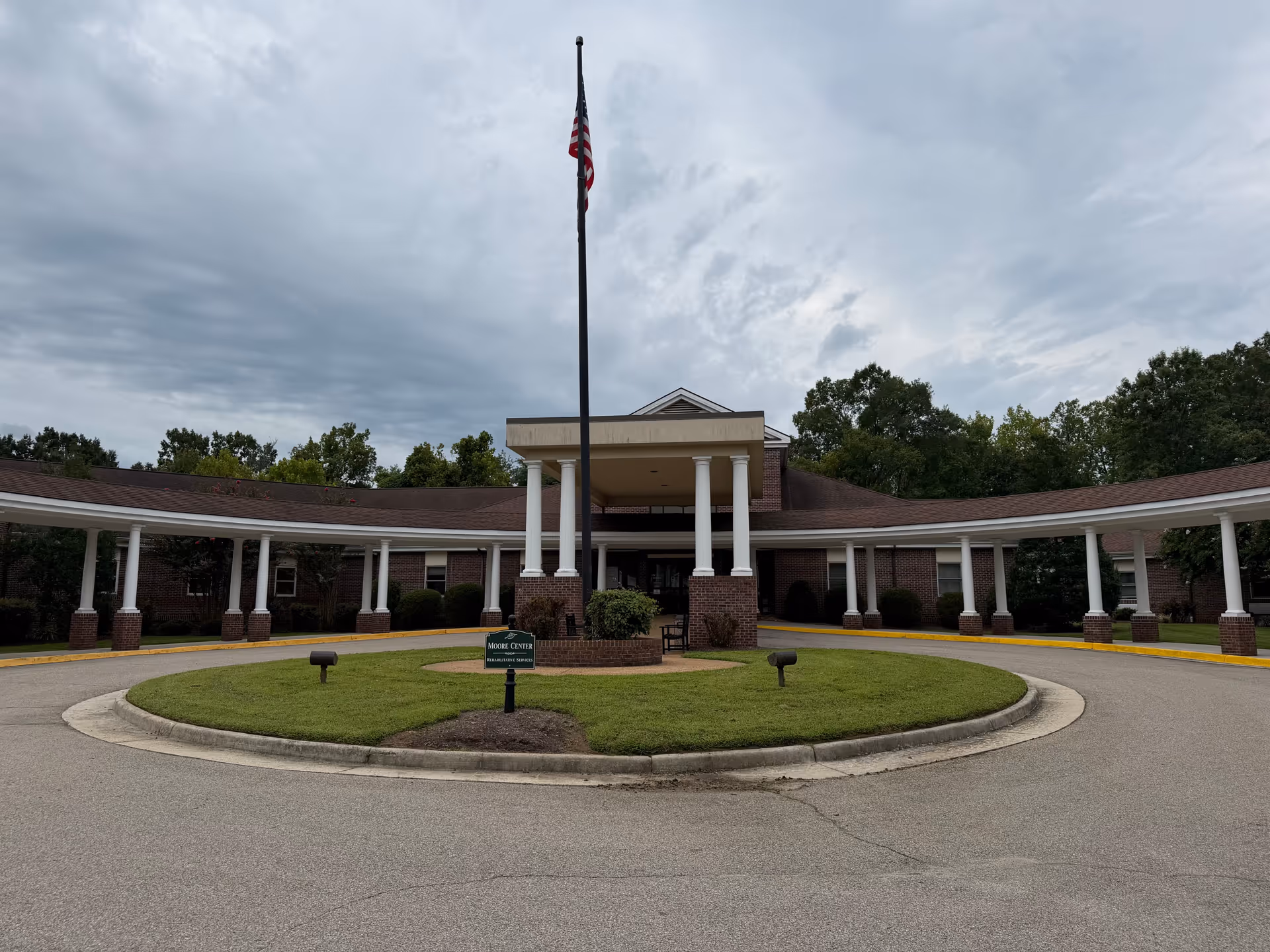 Brick building with a circular driveway, flagpole, and covered entrance under cloudy sky.