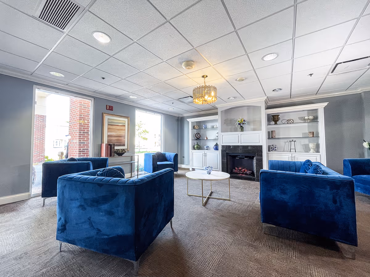 A bright and spacious living room area with four blue velvet armchairs arranged around a round white coffee table. The room features a fireplace with built-in white shelves on either side displaying decorative items. Large windows allow natural light to fill the space, and a modern chandelier hangs from the ceiling.