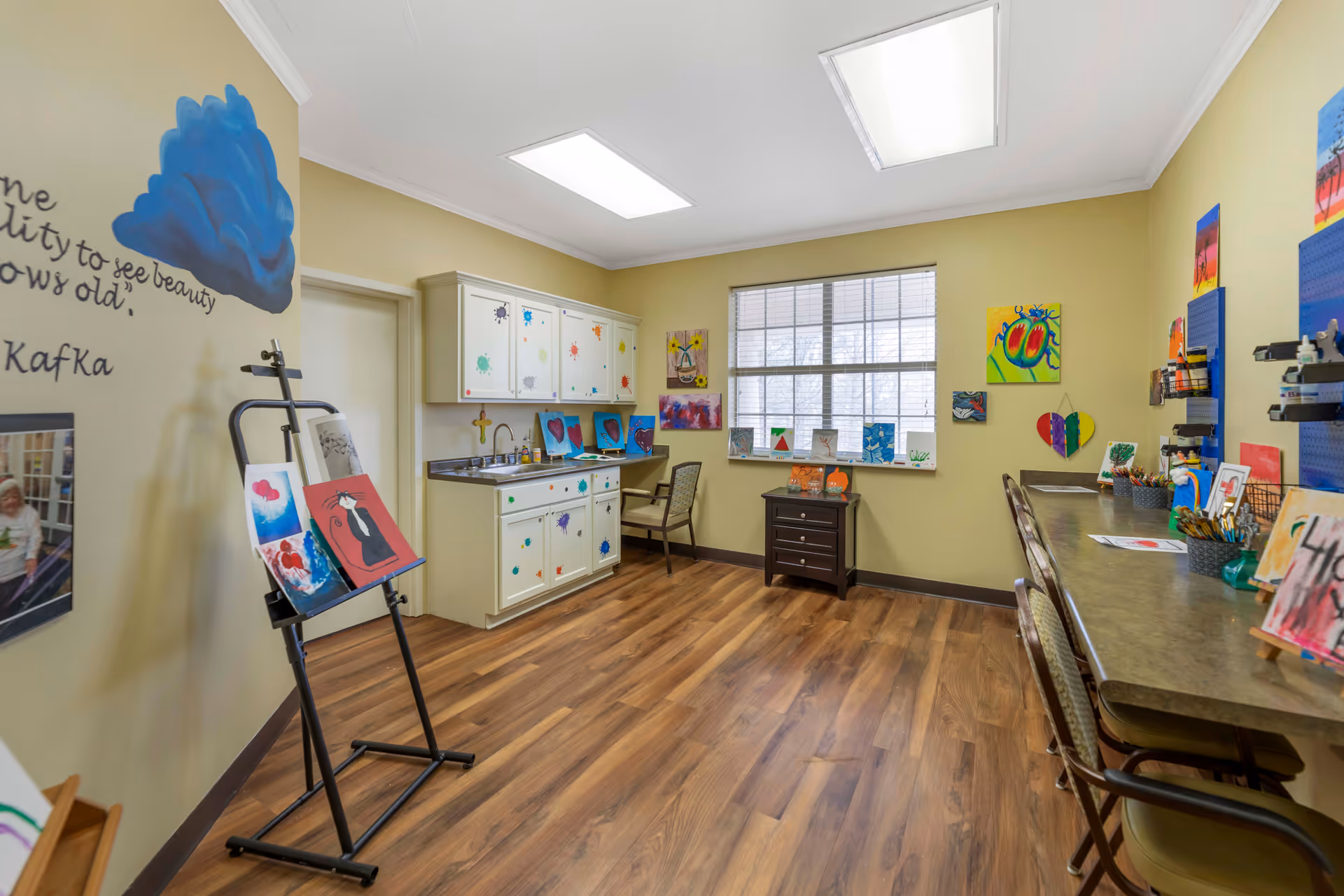 A bright art room with wooden flooring, beige walls, and a large window letting in natural light. The room features a white cabinet with colorful paint splatters, a countertop with a sink, and various colorful paintings displayed on the walls, countertop, and easel. There are chairs along a long desk on the right side, and art supplies are organized on the desk and wall-mounted pegboards.