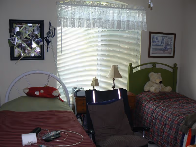 Small bedroom with two twin beds flanking a window, a nightstand with lamps, a teddy bear on one bed, and a chair in front.