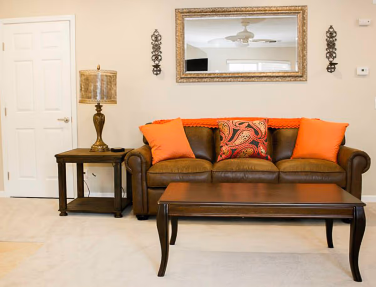 A cozy living room with a brown leather sofa adorned with three orange and patterned cushions. A dark wooden coffee table is in front of the sofa. To the left of the sofa is a matching dark wooden side table with a decorative lamp. Above the sofa hangs a large rectangular mirror with a gold frame, flanked by two ornate wall decorations. The walls are painted beige and there is a white door on the left side.