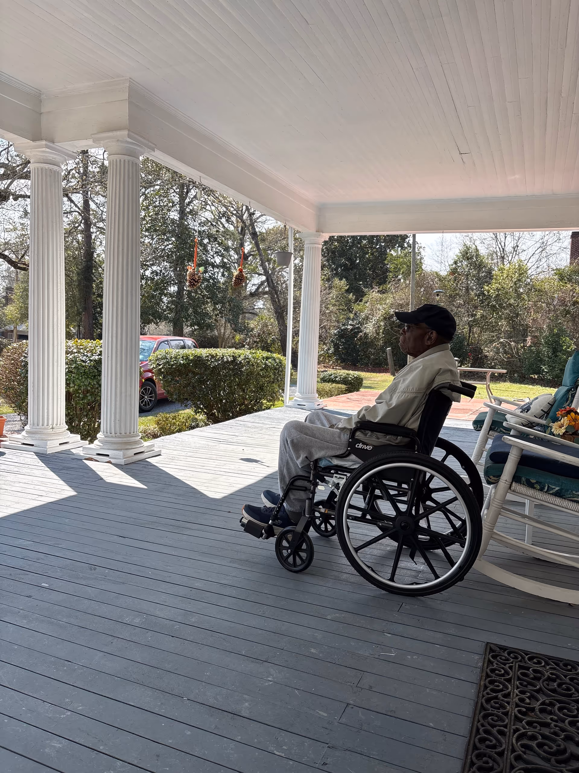 Senior in a wheelchair on a covered porch with columns and outdoor seating.