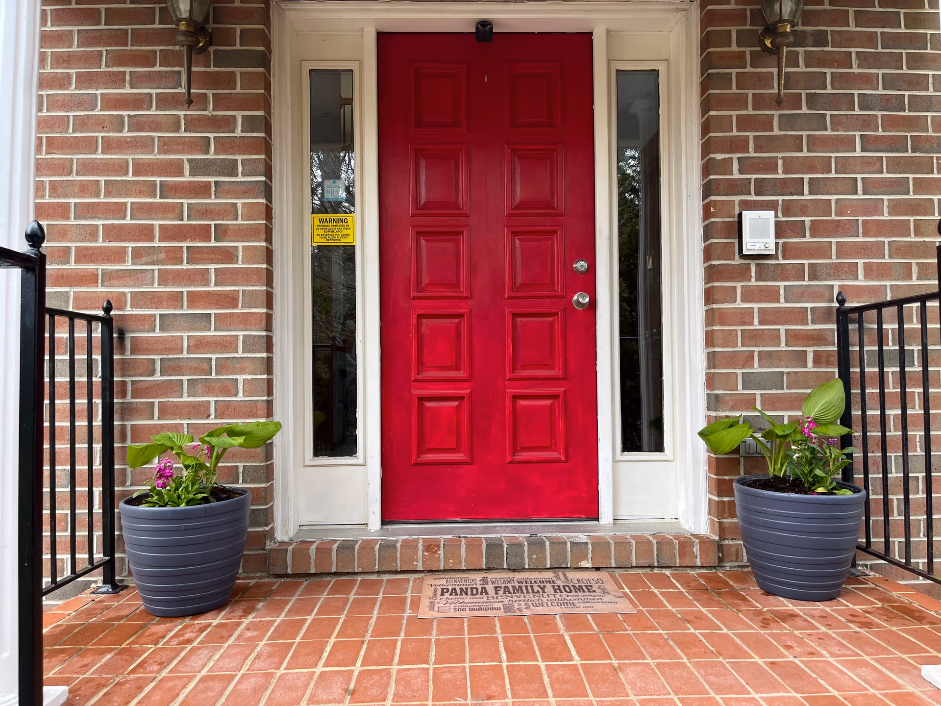 Red front door with brick facade, two potted plants, and a Panda Family Home welcome mat.