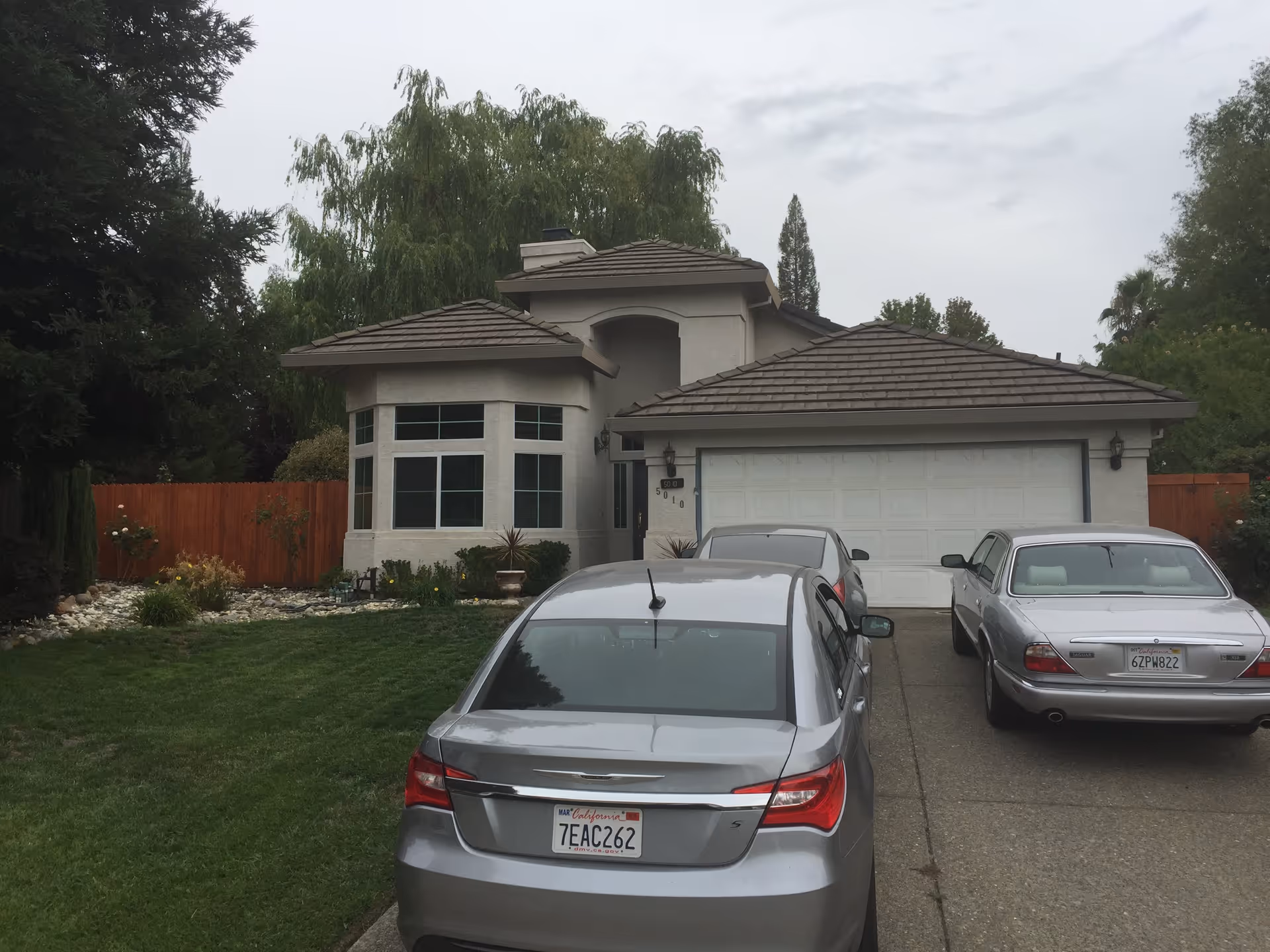 Front exterior view of a single-story house with a two-car garage, a driveway with three parked cars, a well-maintained lawn, and trees and shrubs surrounding the property under a cloudy sky.