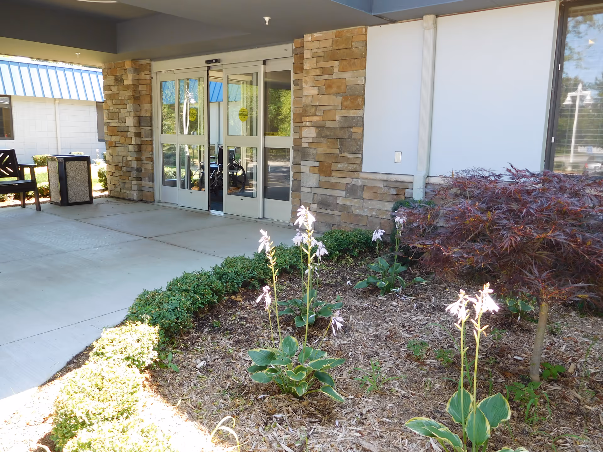 Entrance to a building with automatic sliding glass doors framed by stone pillars. There is a small garden bed with plants and flowers in front of the entrance, and a bench and trash can are visible to the left side under a covered walkway.