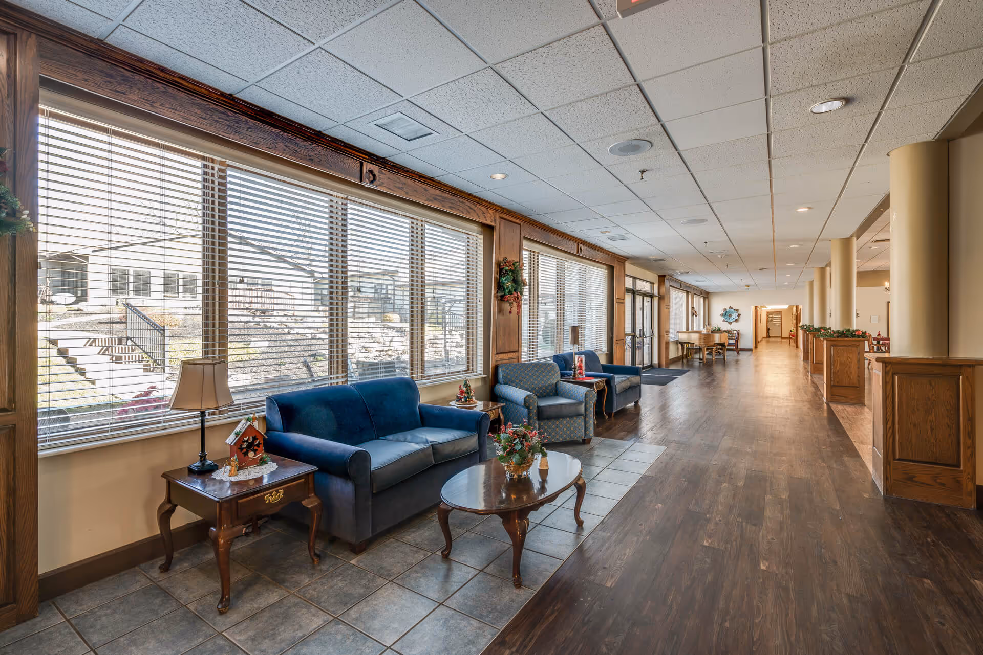 A long hallway in a senior living facility with large windows on the left side letting in natural light. There are blue and patterned armchairs and sofas arranged along the window wall with small wooden tables and decorative items. The floor is a combination of tile near the seating area and wood further down the hallway. The ceiling has recessed lighting and a drop ceiling design. The hallway extends into the distance with more seating and tables visible.