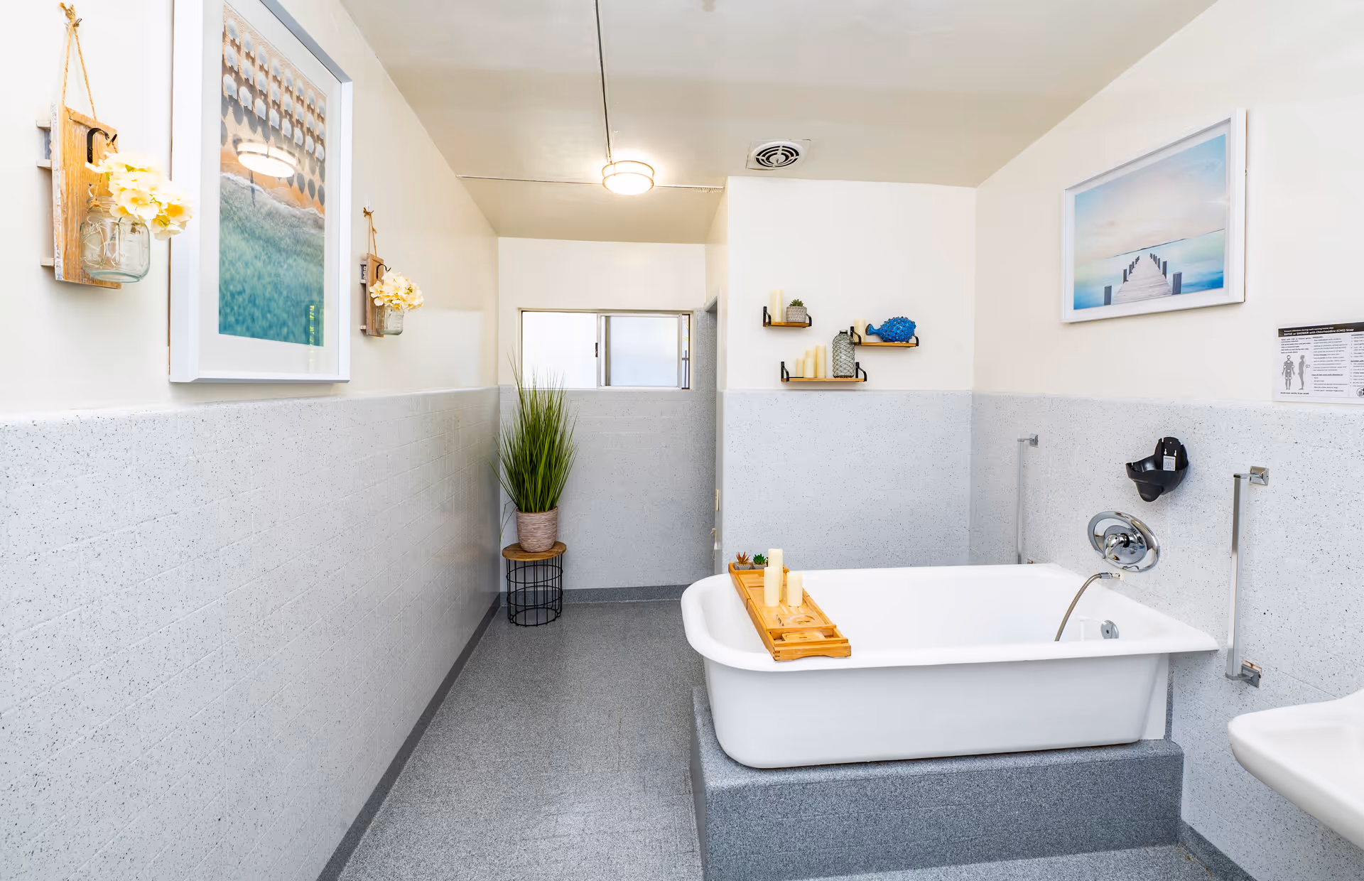 A clean and bright bathroom featuring a white bathtub with a wooden tray holding candles, light gray tiled walls and floor, a small potted plant on a stool, framed pictures on the walls, and shelves with decorative items.
