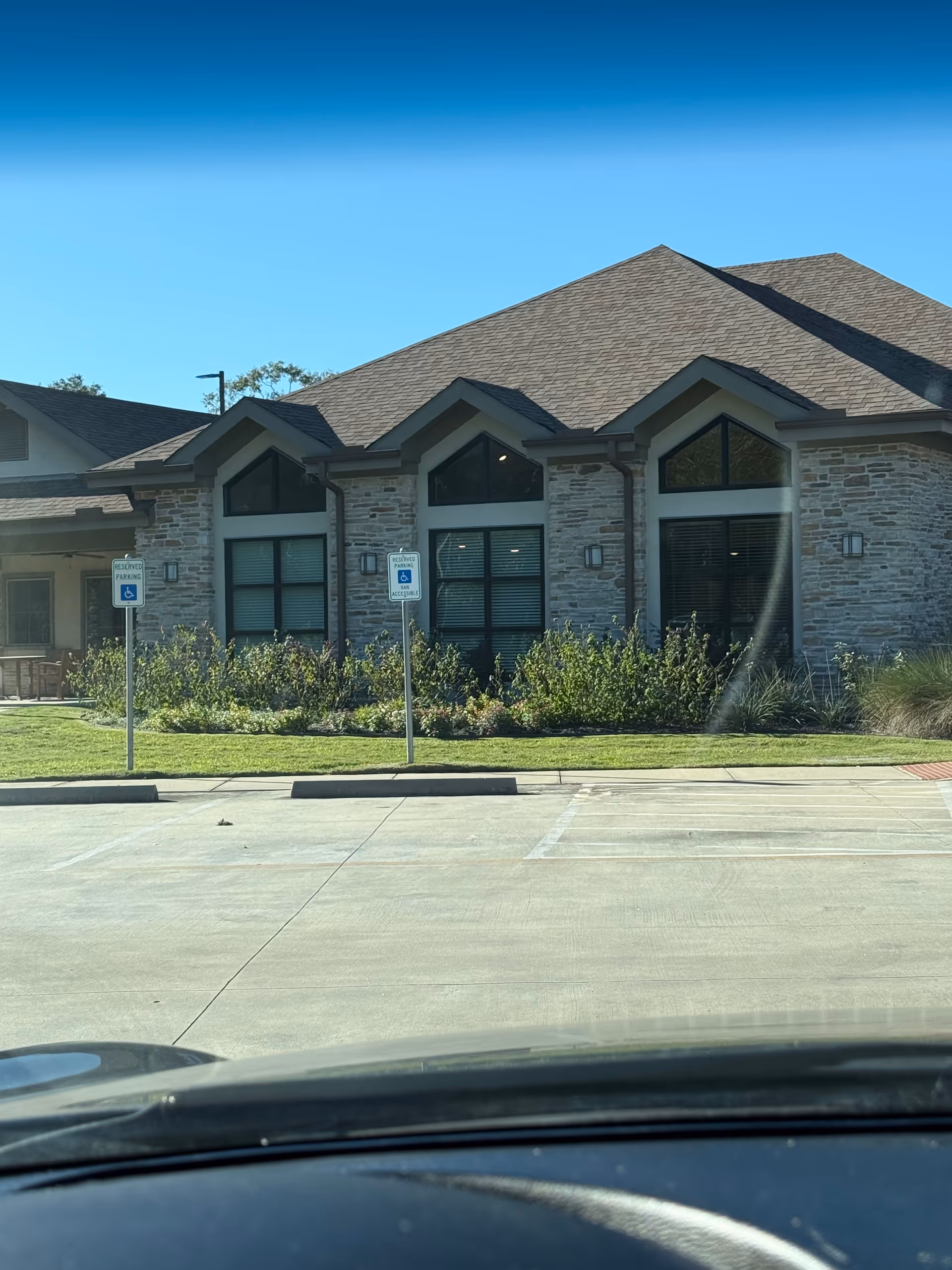 Front view of a stone-faced senior living building with large windows, shrubs, and a parking lot.