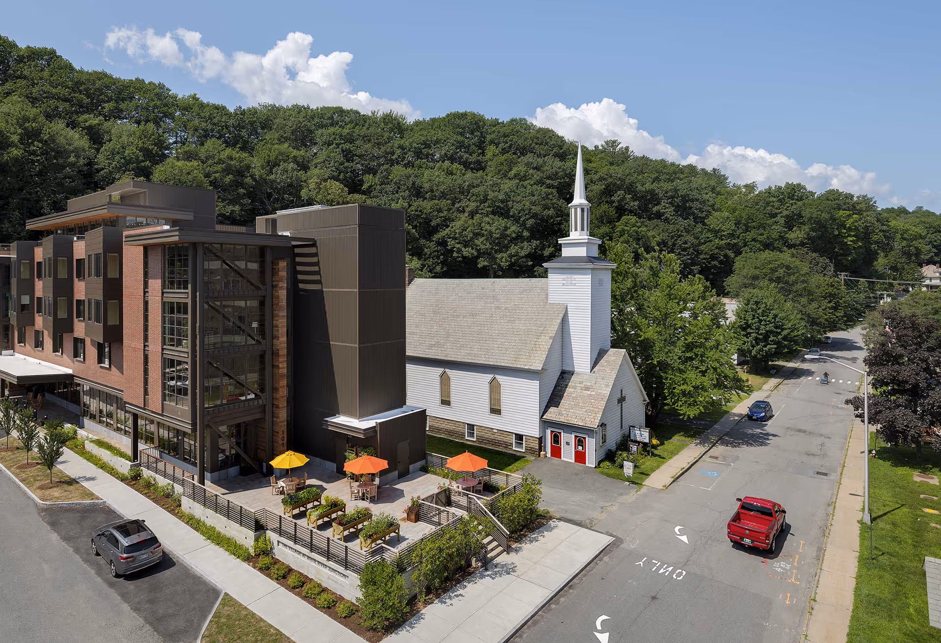 A modern multi-story building with a patio area featuring tables and colorful umbrellas next to a traditional white church with a steeple, surrounded by trees and a street with parked cars.
