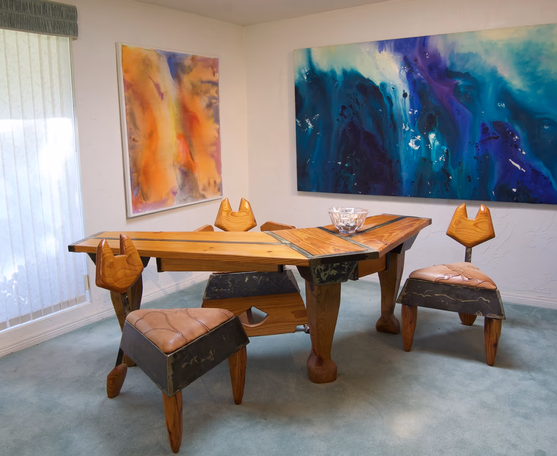 A dining area with a sculptural wooden table and matching carved chairs, colorful abstract paintings on the walls, and a large window with vertical blinds.