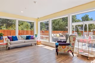 Sunlit living room with large windows overlooking a fenced patio, featuring a sofa, colorful armchair, and hardwood floors.