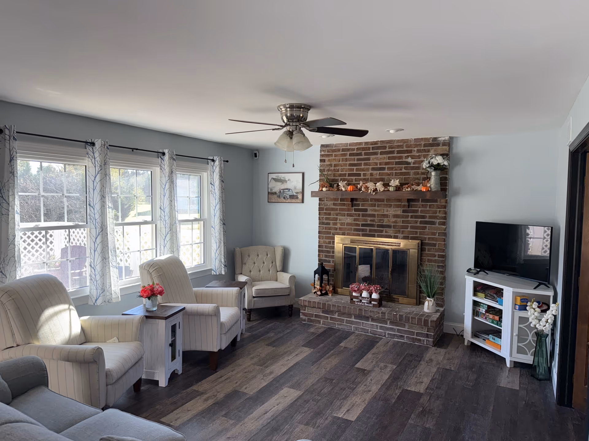 Cozy living room with brick fireplace, armchairs, windows, and a TV in a senior facility.