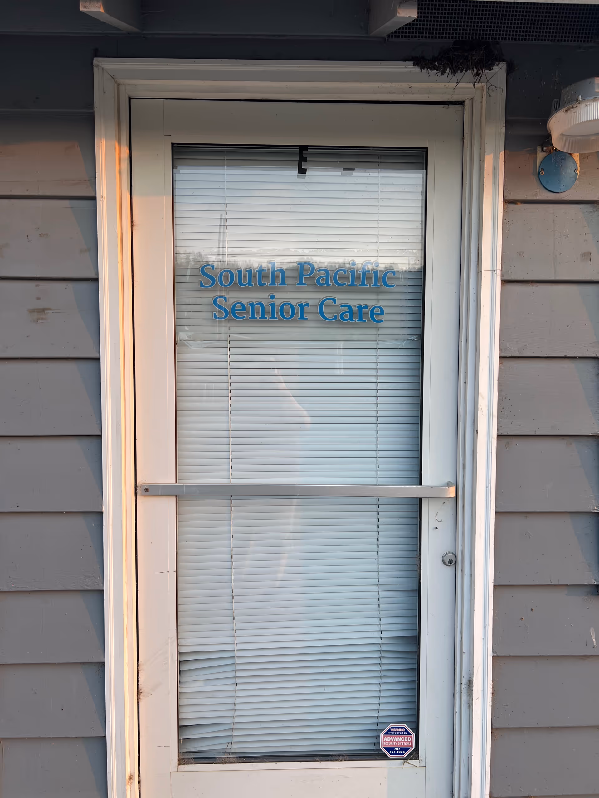 Exterior door labeled South Pacific Senior Care with blinds inside and siding.