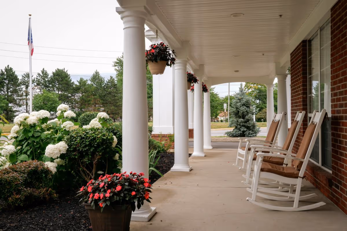 Covered porch with white columns and rocking chairs beside potted and hanging flowers at the front of a brick building.