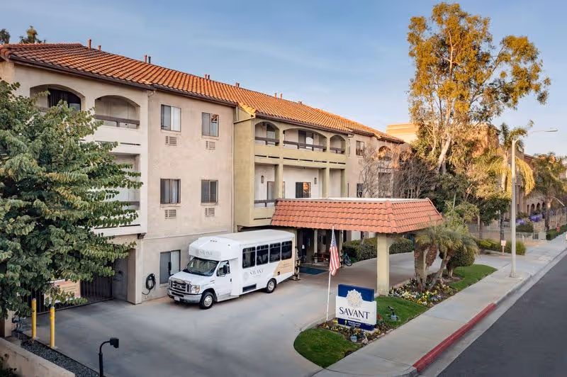 Front entrance of a multi-story senior living facility with a covered porte-cochere, a Savant shuttle van, and a Savant sign by the driveway.