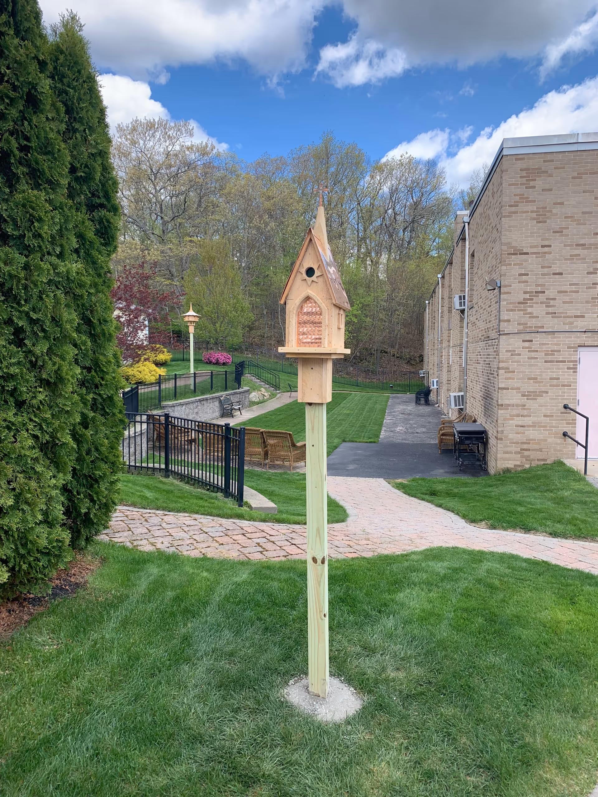 A small wooden birdhouse mounted on a tall wooden post in a grassy area near a brick building. There is a paved walkway, black metal fencing, benches, and trees in the background under a partly cloudy blue sky.
