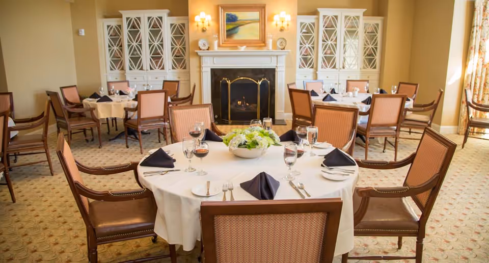 A dining room with round tables covered in white tablecloths, set with plates, glasses, silverware, and black napkins. The room features wooden chairs with cushioned seats, a fireplace with a decorative screen, built-in cabinets with glass doors, wall sconces, and a painting above the fireplace. The carpet has a subtle pattern, and natural light comes through a window with floral curtains.