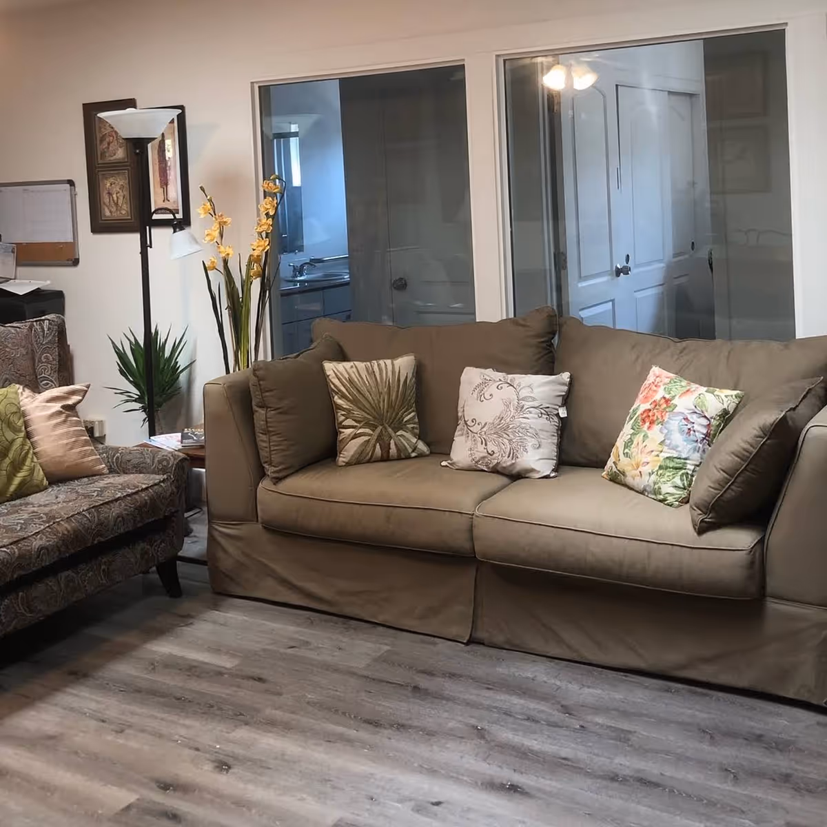 A cozy living room area with a beige sofa adorned with three decorative pillows featuring floral and leaf patterns. To the left, there is a patterned armchair with two pillows, a tall floor lamp, and two potted plants. Behind the sofa, there are two large windows showing a bathroom with a sink and a door. The floor is covered with light wood laminate.