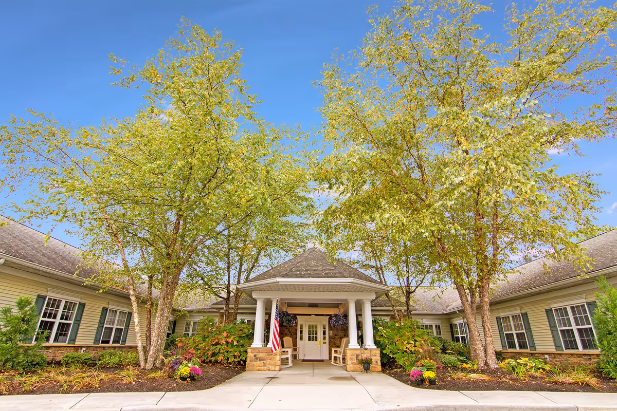 Entrance of a senior living facility with a covered porch supported by white columns, surrounded by trees and landscaped plants. The building has beige siding with green shutters and a stone base. An American flag is displayed near the entrance under a clear blue sky.