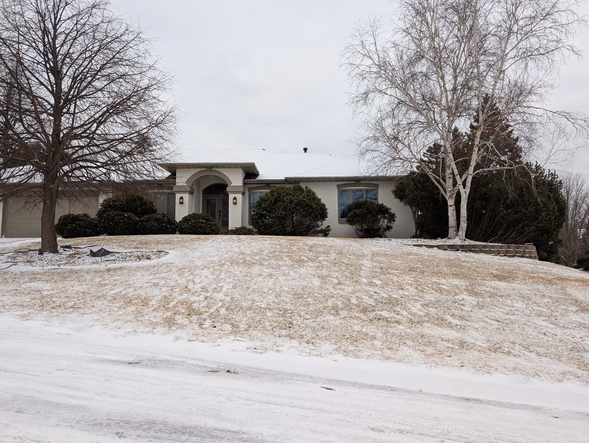 Front exterior of a single-story house with an arched entry, garage, bare trees and a light dusting of snow on the lawn.