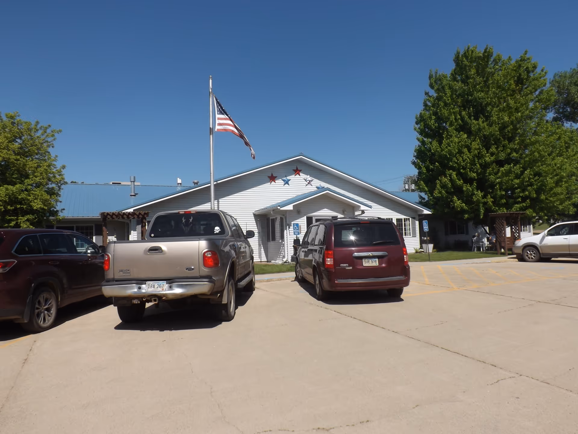 Single-story assisted living building with an American flag in front and several parked cars in the lot.
