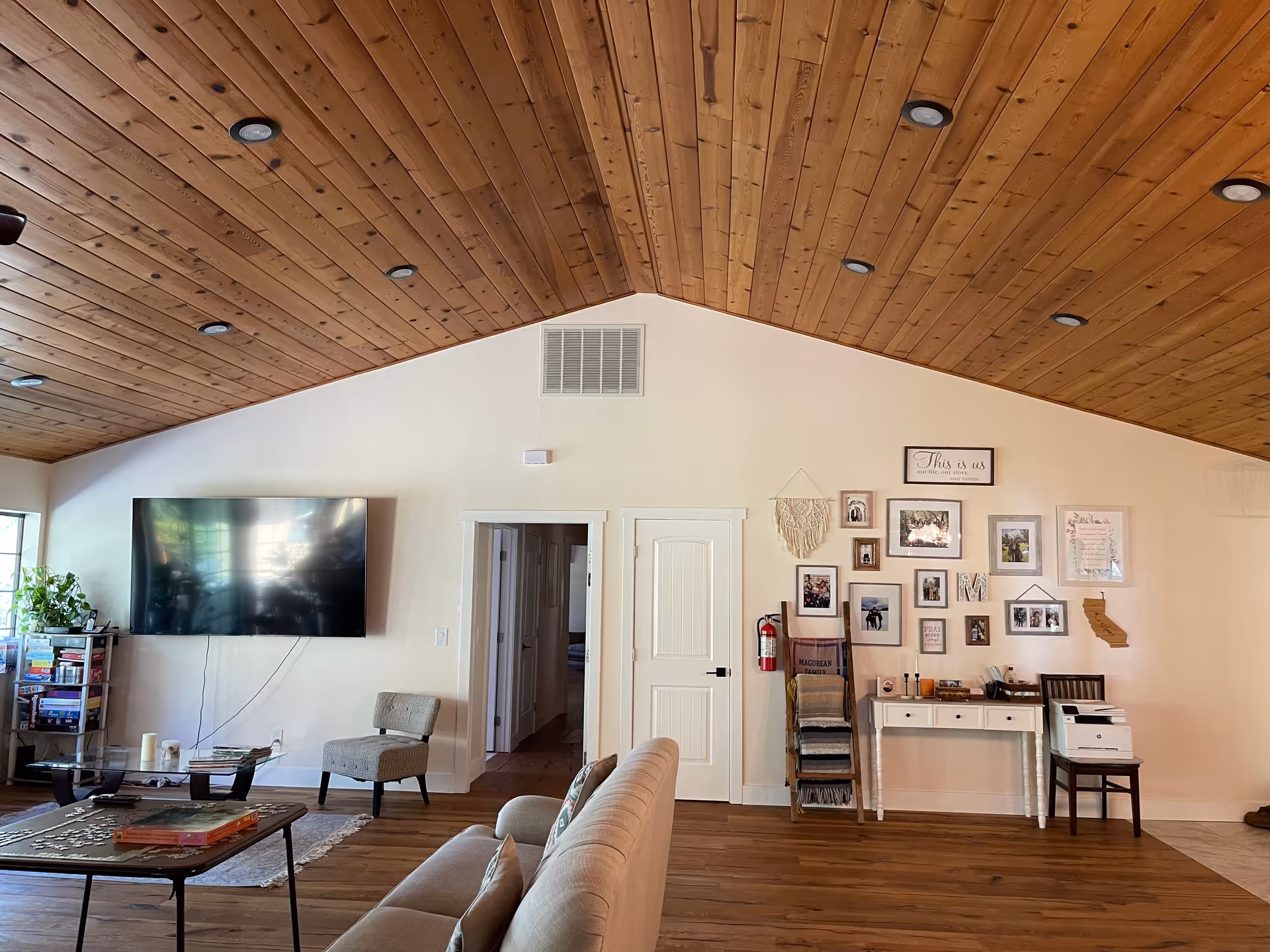 Spacious living room with a wood-paneled vaulted ceiling, mounted flat-screen TV, sofa, chairs, coffee table and a gallery wall above a console table.