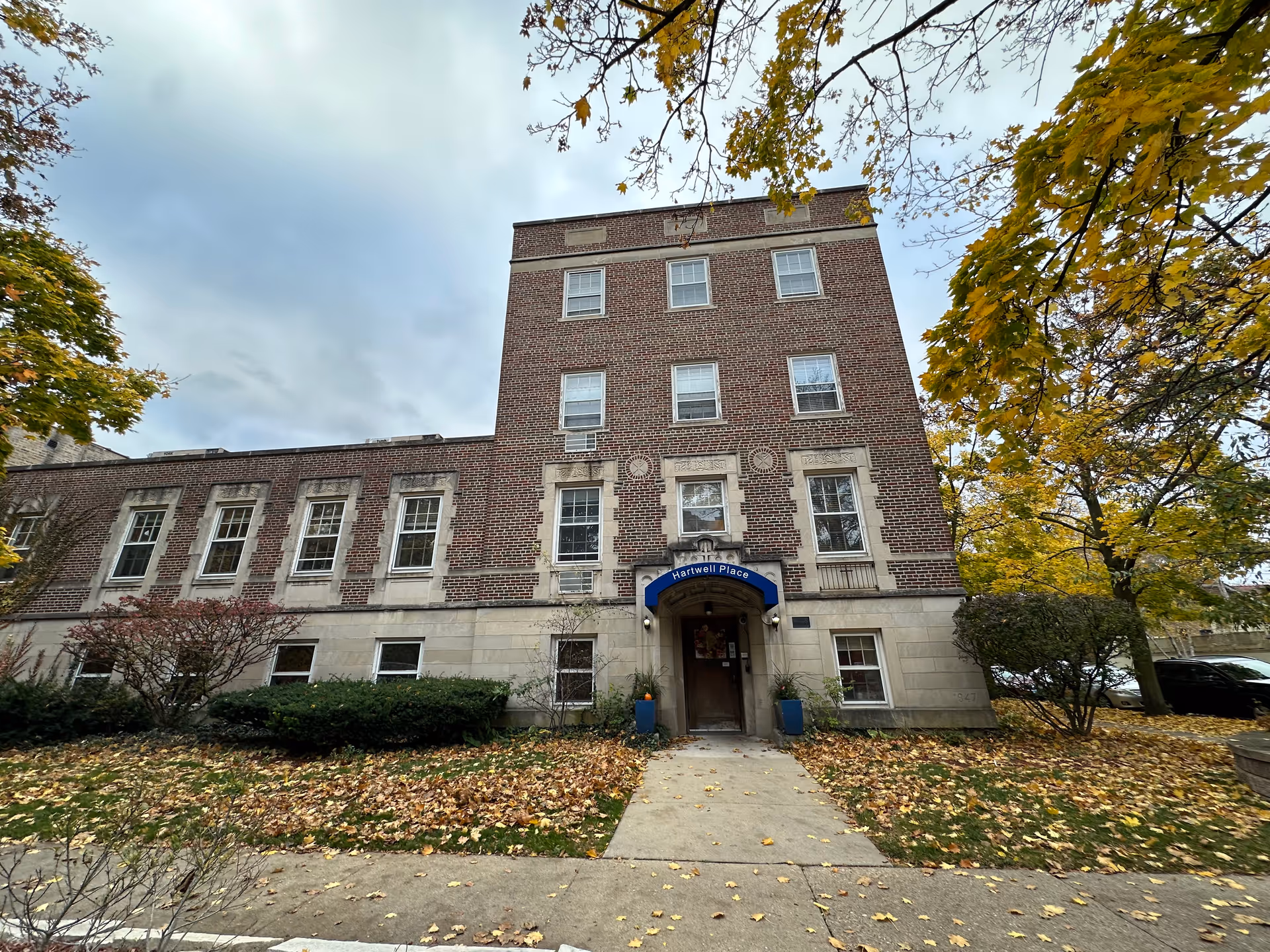 Exterior view of a multi-story brick building with a blue awning above the entrance that reads 'Hartwell Place'. The building is surrounded by trees with autumn leaves and a sidewalk leading to the entrance.