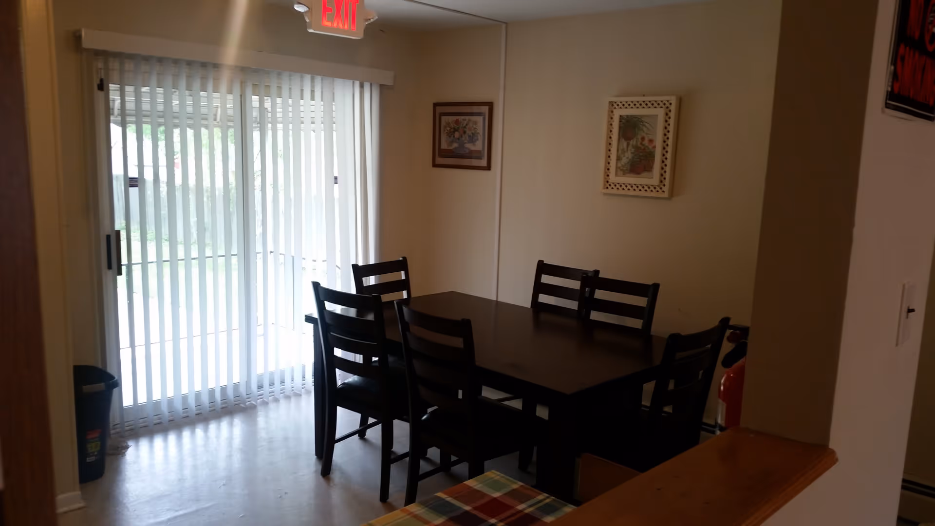 A dining area with a dark wooden table and six matching chairs. The room has beige walls with two framed floral pictures hanging. There is a large sliding glass door with vertical blinds letting in natural light. An exit sign is visible above the door, and a fire extinguisher is partially visible near the corner.