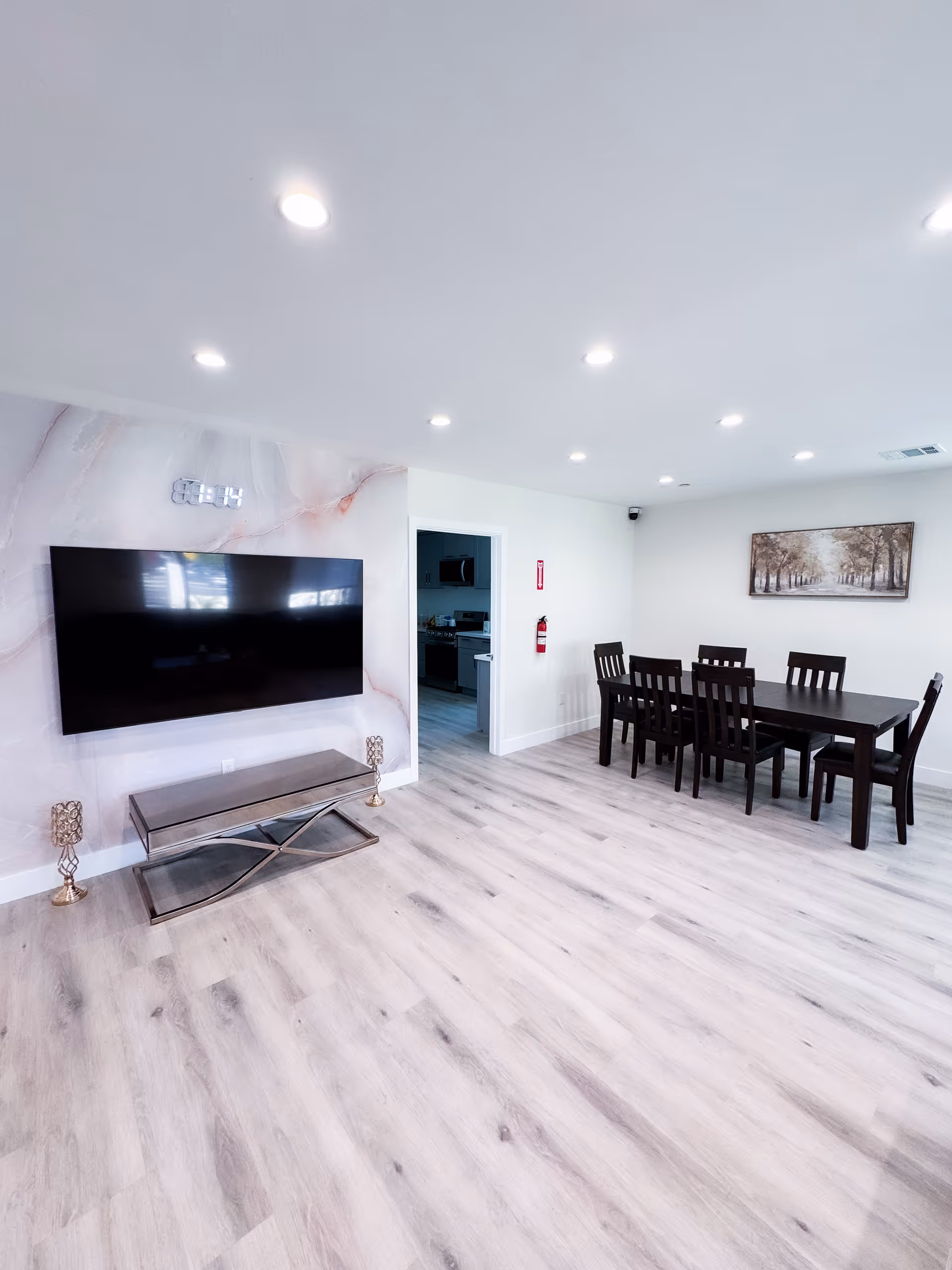 A bright, modern interior room with light wood flooring and white walls. On the left wall, a large flat-screen TV is mounted above a glass and metal console table with two decorative candle holders on the floor. To the right, there is a dark wooden dining table with six matching chairs. A framed picture of a tree-lined path hangs on the far wall. A doorway leads to a kitchen area in the background.