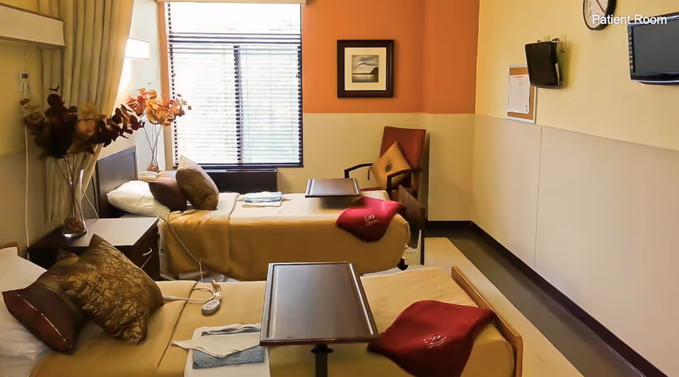 A patient room with two hospital beds, each with beige bedding and decorative pillows. Each bed has a small overbed table and a folded red blanket. There is a window with blinds letting in natural light, a chair with a cushion, a wall-mounted TV, and a clock on the wall. A vase with dried flowers is on a bedside table.
