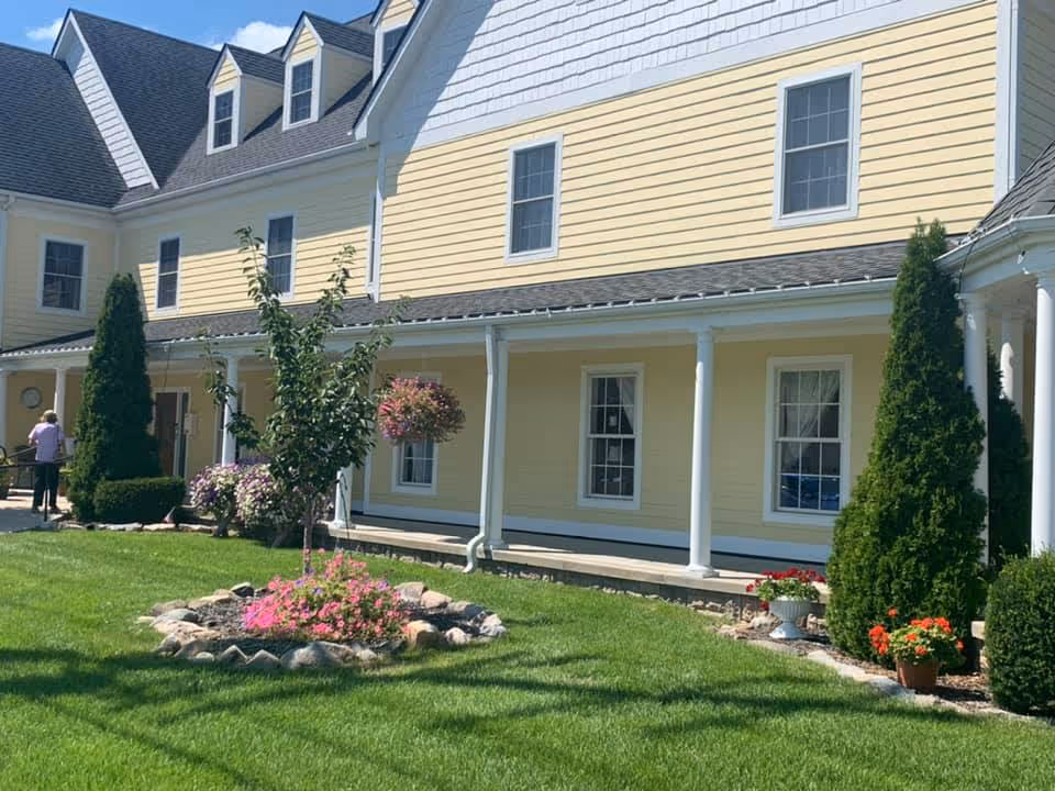 Front exterior of a yellow two-story building with a covered porch, white columns, manicured lawn, and flowerbeds.