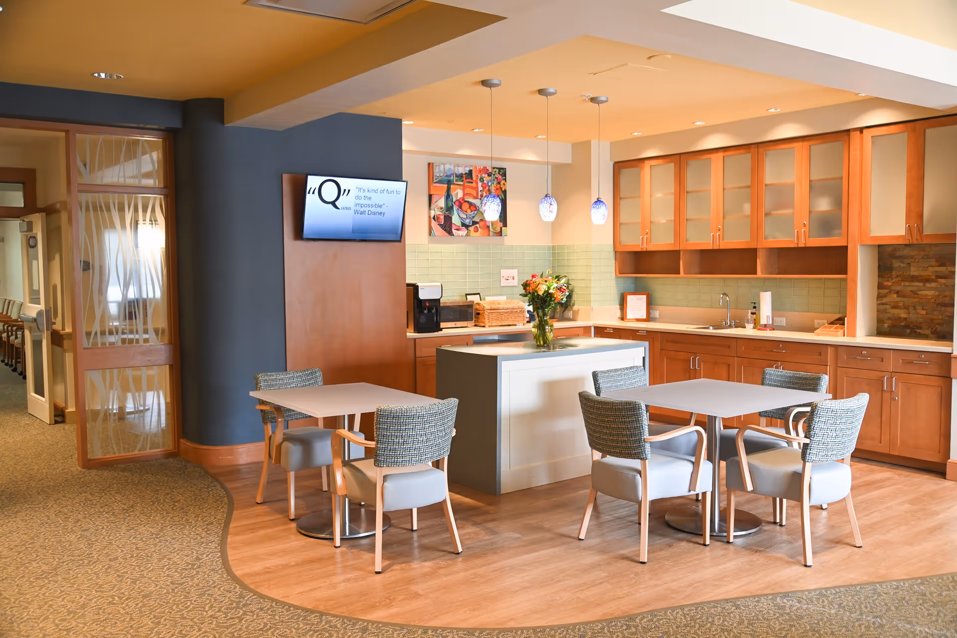 A bright and modern kitchen and dining area in a senior living facility with wooden cabinets, a central island, and two small square tables each surrounded by four cushioned chairs. The space features pendant lights, a wall-mounted TV displaying a quote, and a vase with flowers on the island. The floor transitions from carpet to wood, and a glass door with a decorative pattern is visible on the left.