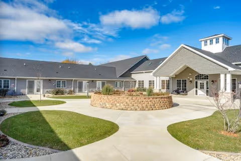 Exterior view of a single-story assisted living facility building with a covered entrance, a circular raised flower bed in the center of a paved courtyard, green grass areas, and a partly cloudy blue sky.