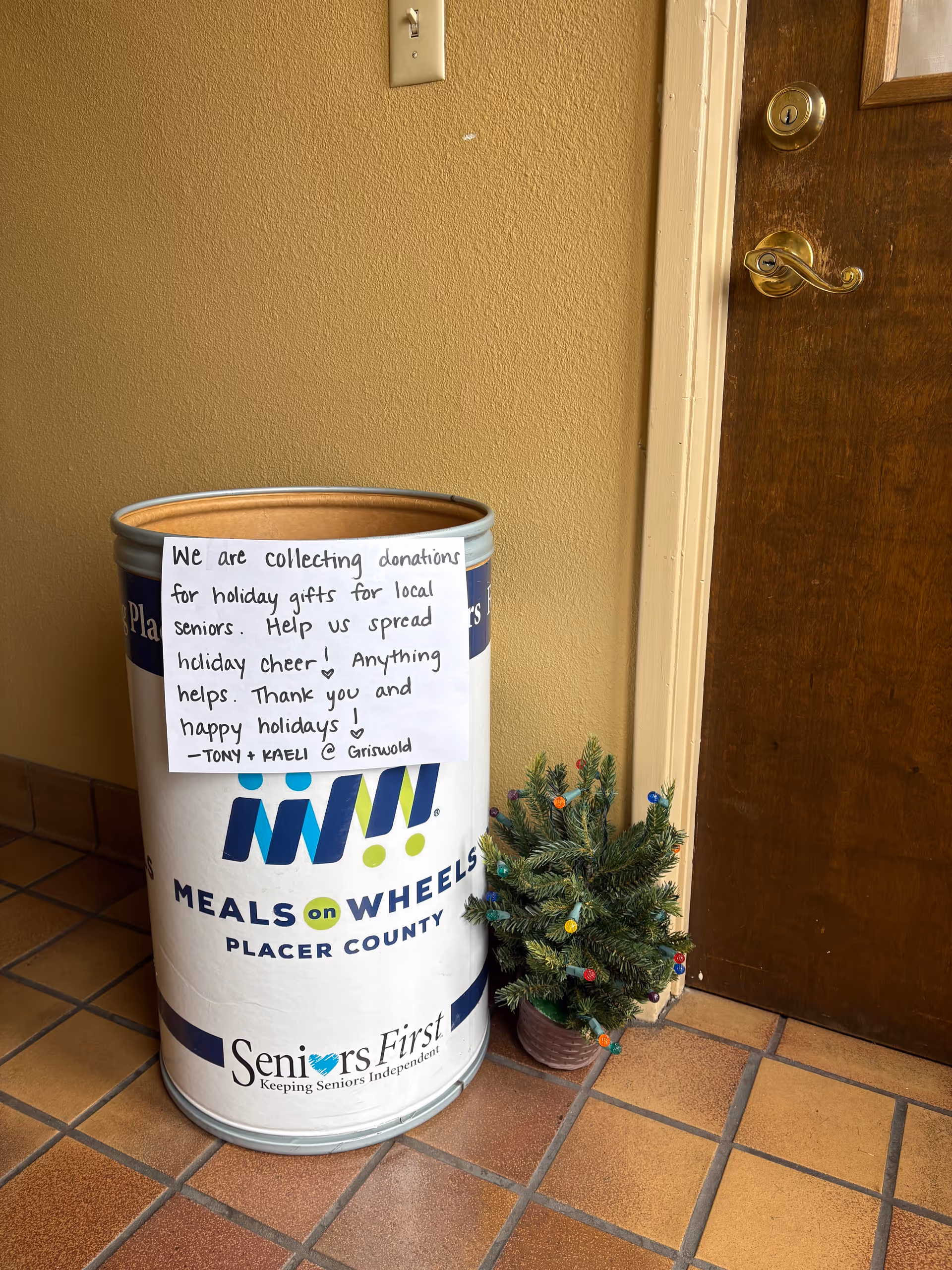 Donation bin for Meals on Wheels next to a small Christmas tree by a door.