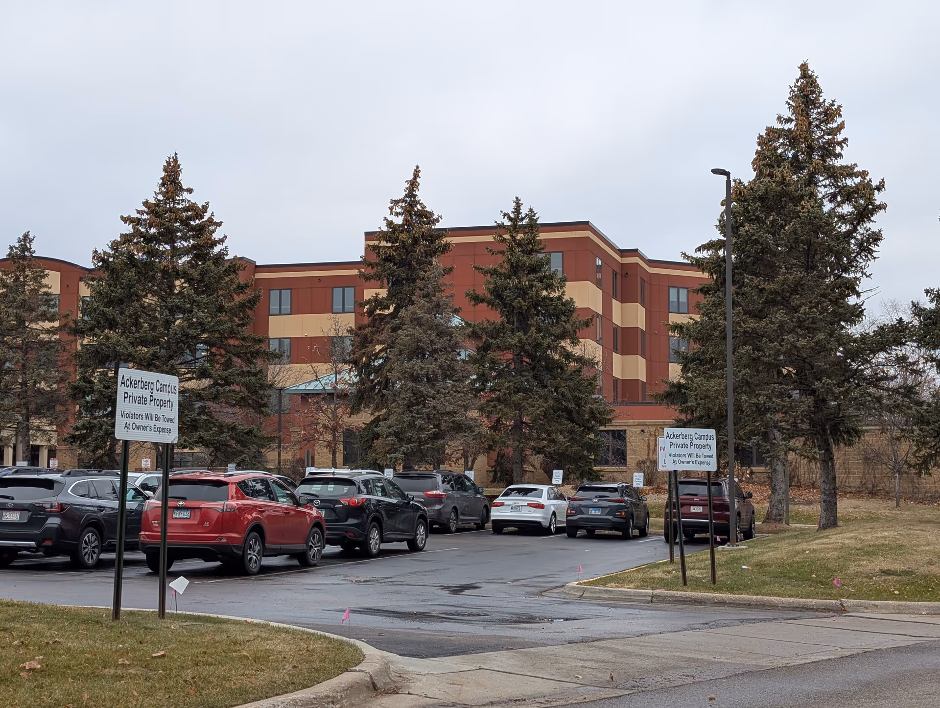 Exterior view of a multi-story senior living building with parked cars, evergreen trees, and private property signs in the foreground.