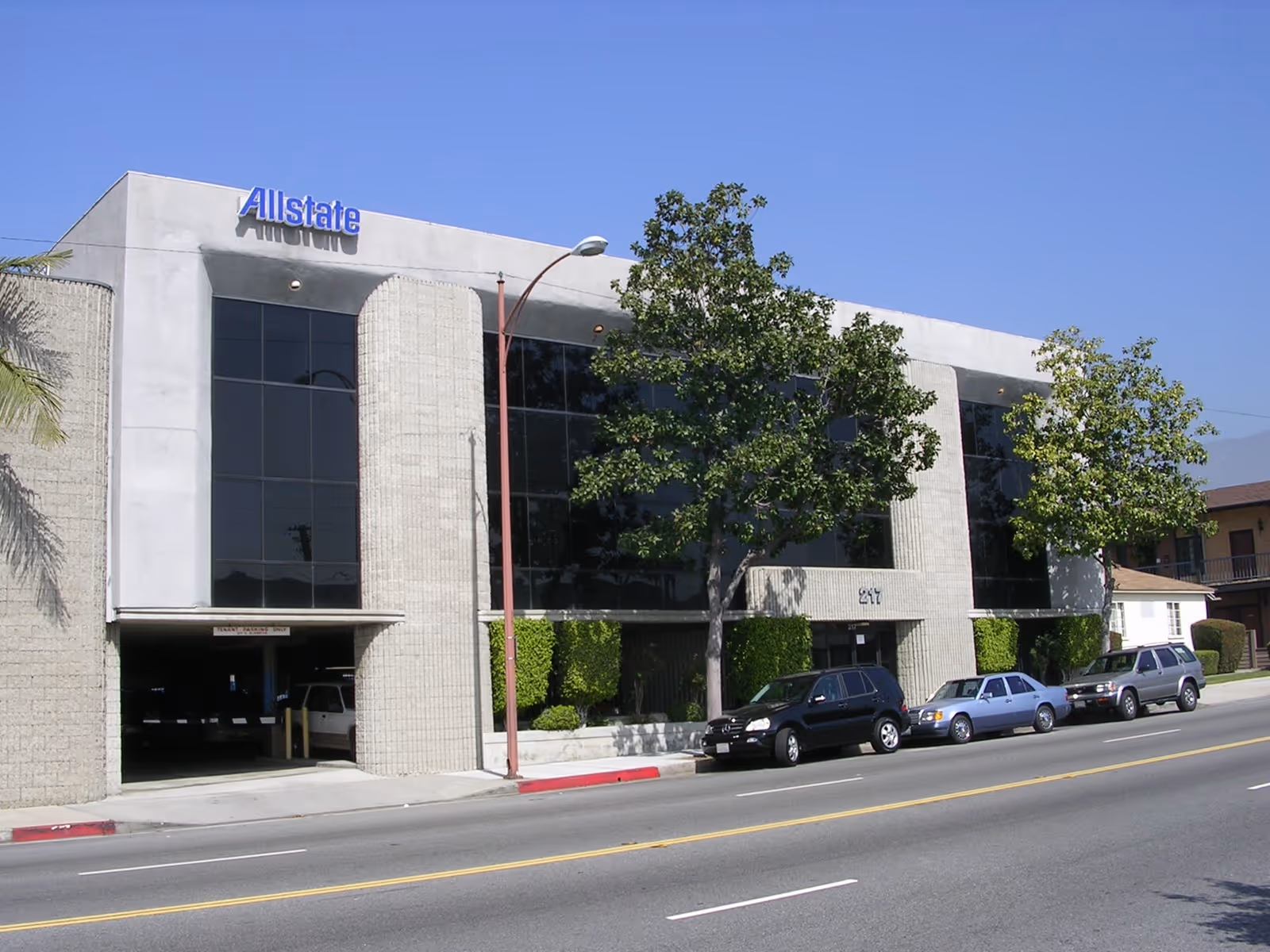 A modern, two-story building with large glass windows, an Allstate sign, trees, and parked cars.