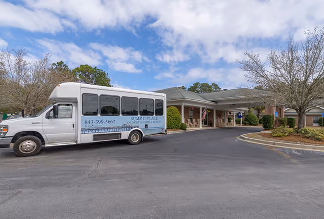 A white shuttle bus parked in front of a single-story brick building with a covered entrance. The bus has the text 'SUMMIT PLACE OF NORTH MYRTLE BEACH' and a phone number on its side. The building has rocking chairs on the porch and an American flag near the entrance. Trees and shrubs surround the area under a partly cloudy sky.