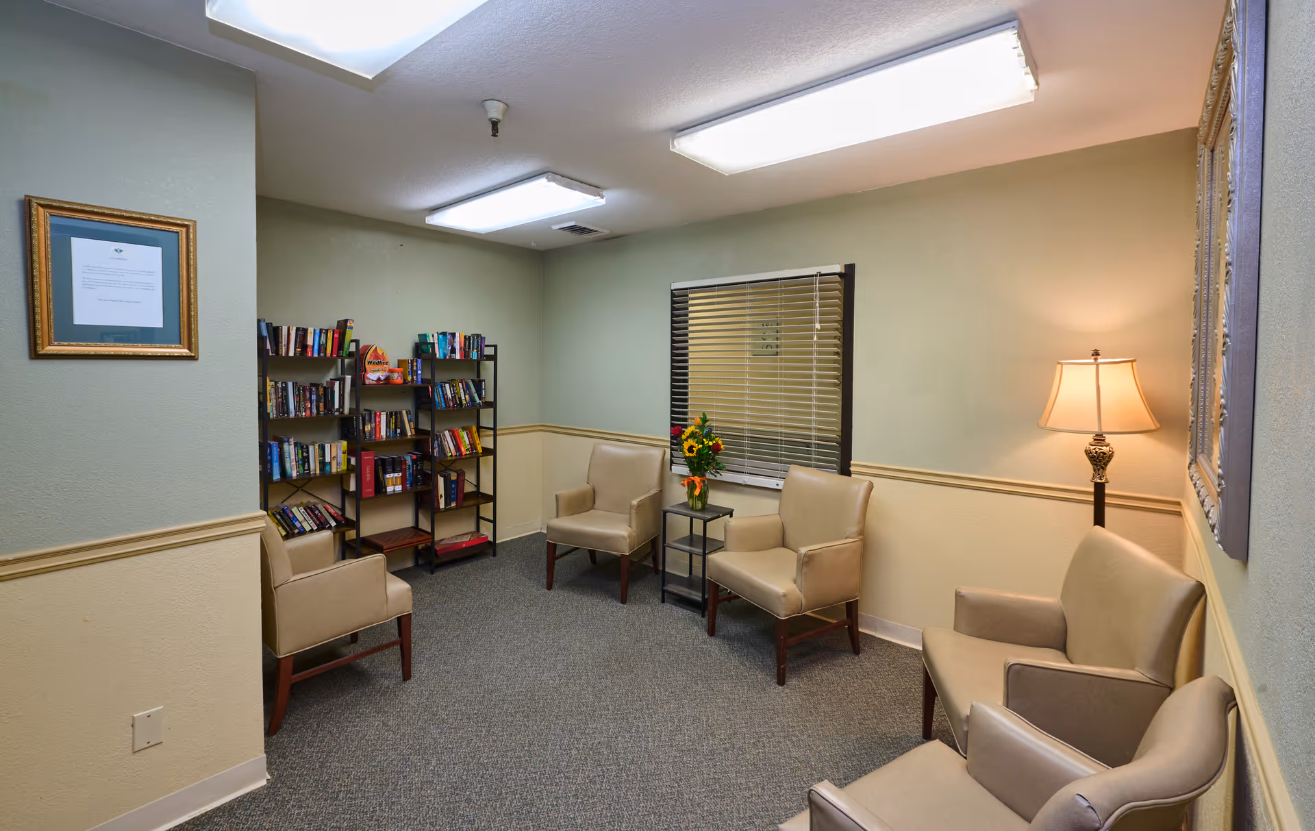 Small sitting room with several upholstered chairs, a bookshelf, side table with flowers, and a floor lamp.