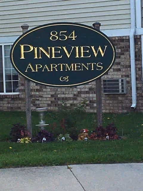 Green oval sign reading '854 Pineview Apartments' mounted on posts in front of a brick apartment building.