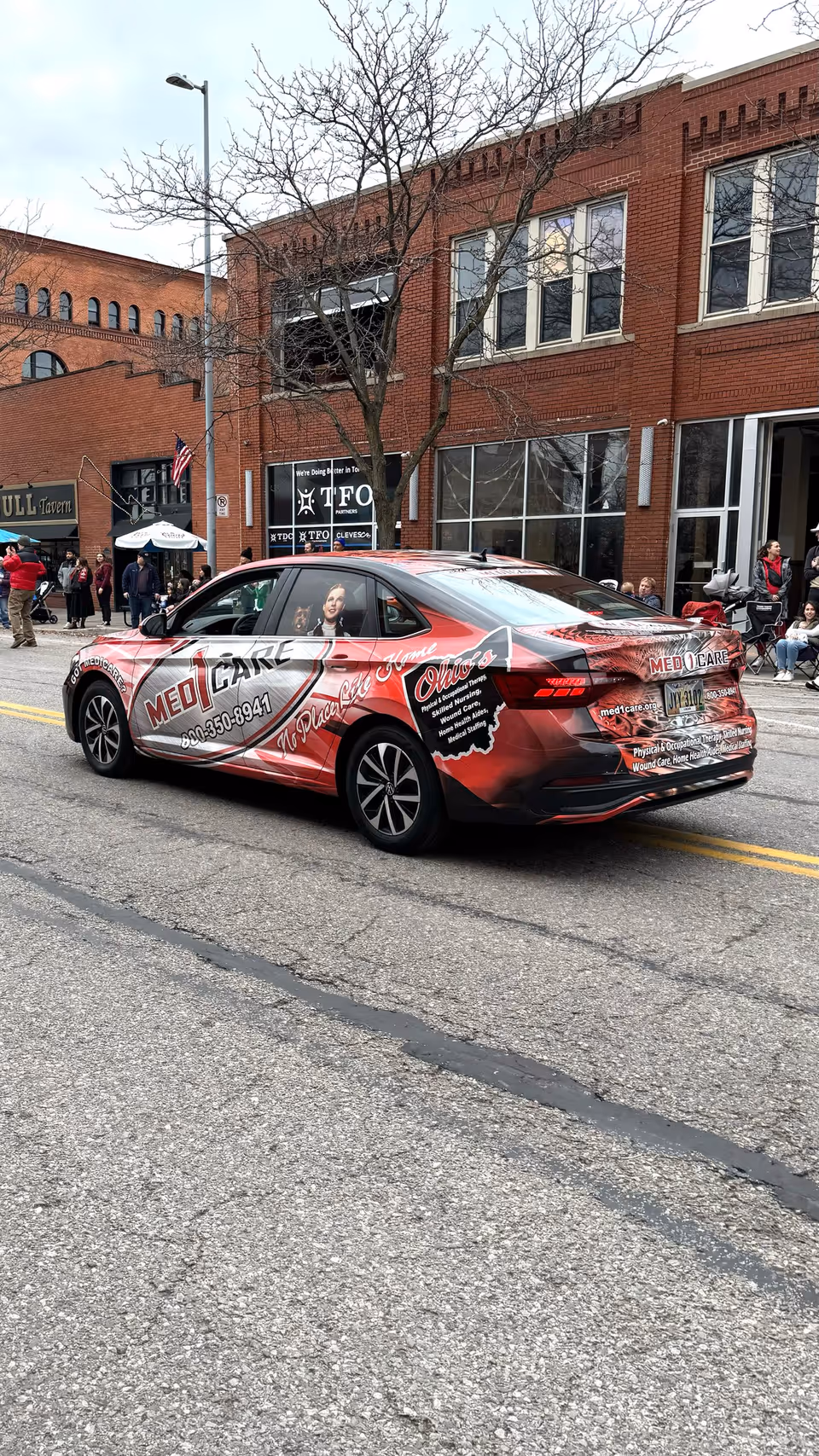 Red promotional Med1Care car drives past brick storefronts on a city street.