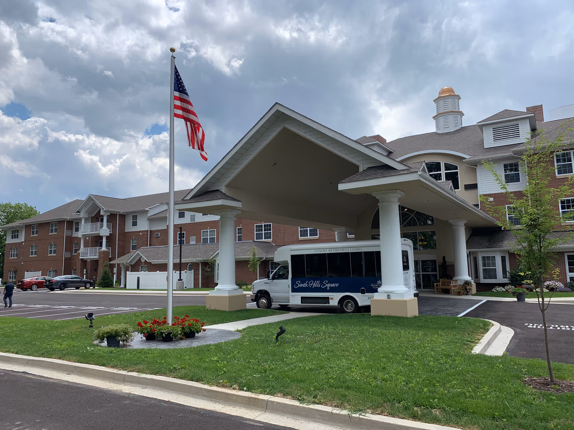 Exterior view of South Hills Square Retirement Resort showing a large covered entrance with white pillars, an American flag on a flagpole, a shuttle bus parked under the entrance, and a well-maintained lawn with flower pots. The building is a multi-story brick and siding structure with multiple windows and a cupola on the roof under a partly cloudy sky.