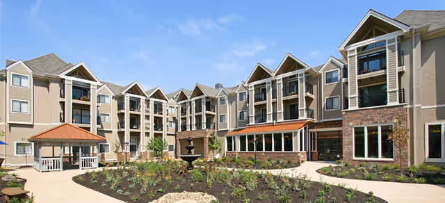 Exterior view of a multi-story senior living facility with beige and brown siding, multiple balconies, large windows, and a landscaped courtyard featuring a gazebo and a central water fountain under a clear blue sky.