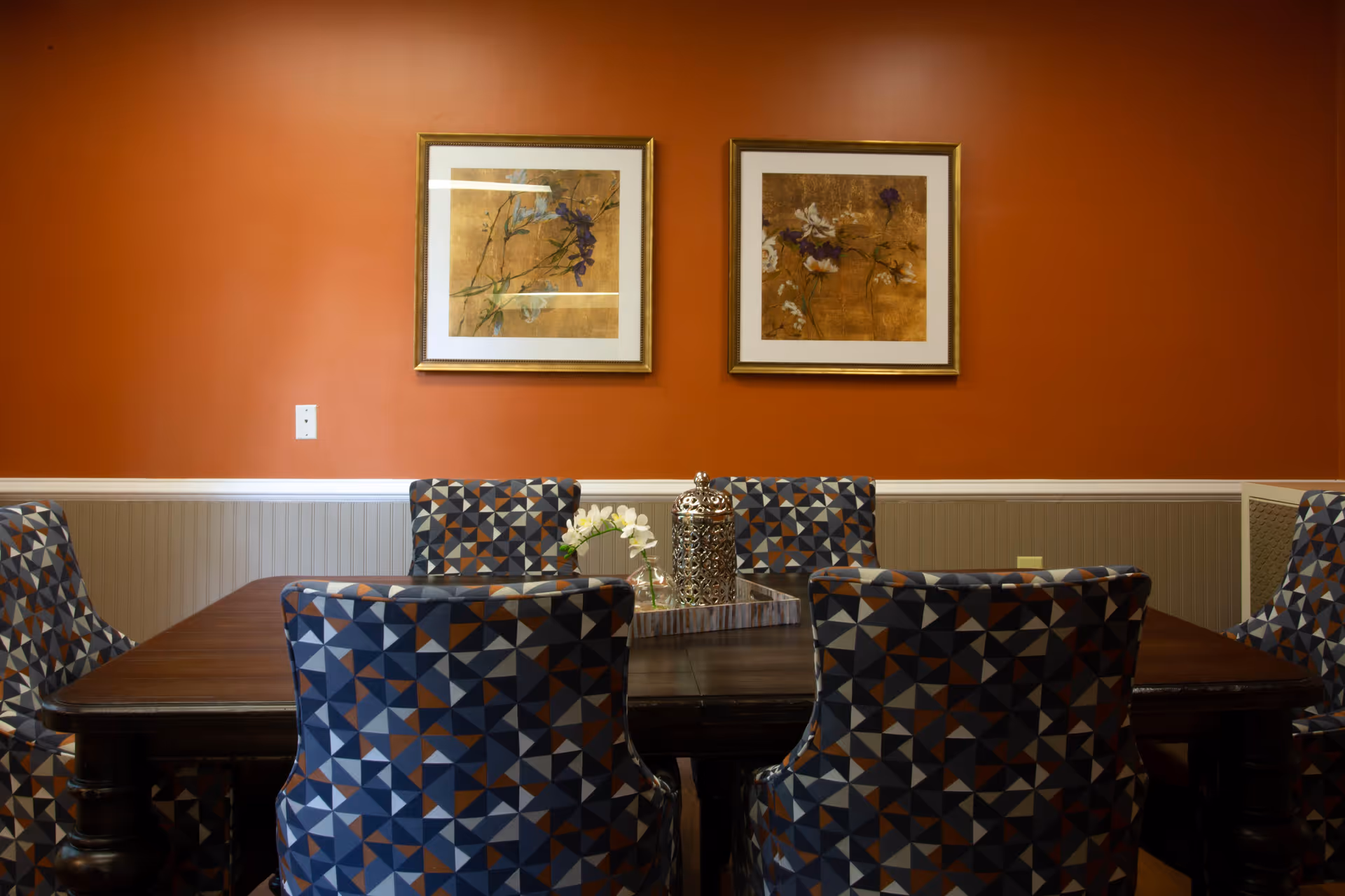 Dining room with wooden table, geometric chairs, orange walls, and framed art.