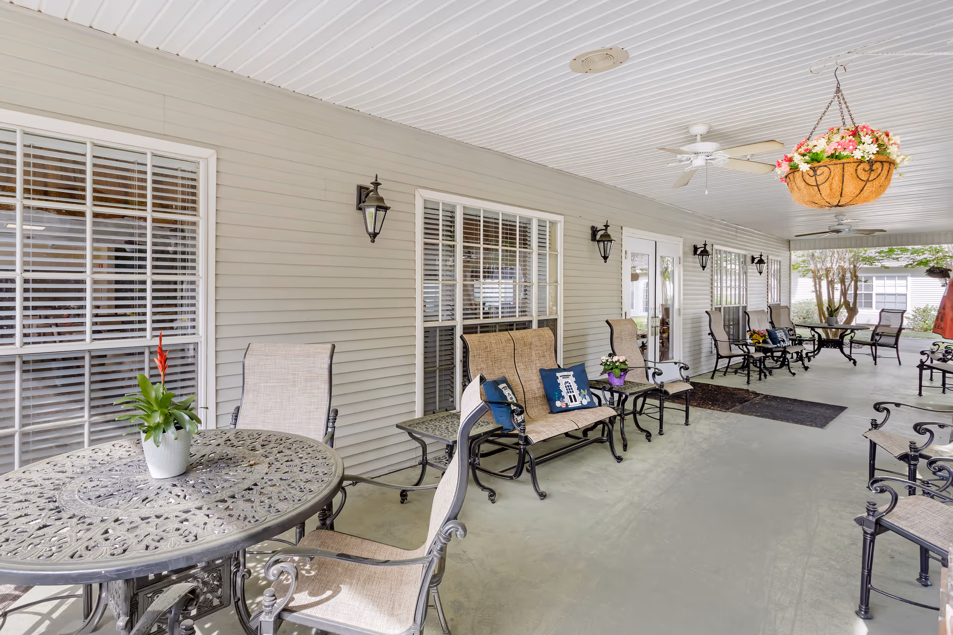Covered outdoor patio with metal tables, cushioned chairs, hanging flower basket, and glass doors.
